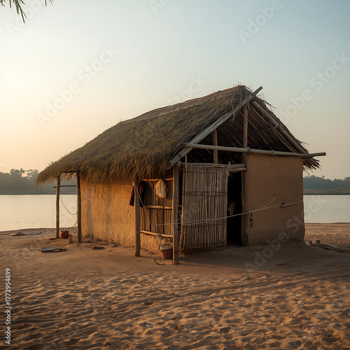 A small, handmade riverside hut features a straw roof and clay walls, constructed of local materials. It stands by a calm river at sunset.