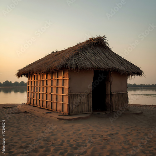 A small, handmade riverside hut features a straw roof and clay walls, constructed of local materials. It stands by a calm river at sunset.