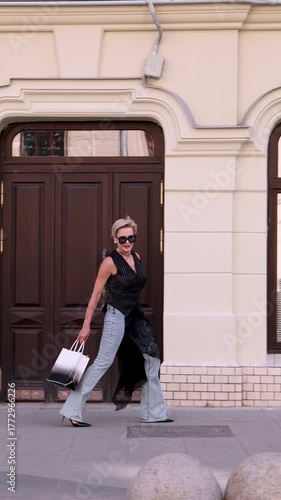 Fashionable Mature woman walking outdoors, wearing sleeveless black polka dot blouse, light blue jeans, black shoes and carrying stylish handbag