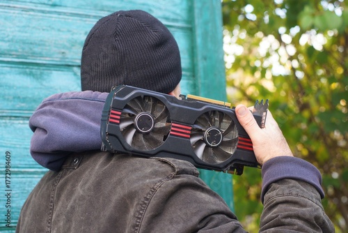 One black modern iron with two round coolers powerful electric graphic gaming video card for computer in hand on shoulder of man in hat and jacket near green wooden wall