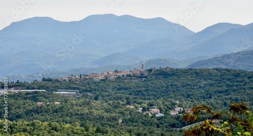 Wide panoramic view of a densely populated valley and town Kastav, Croatia, surrounded by rolling, forested mountains under a hazy sky.