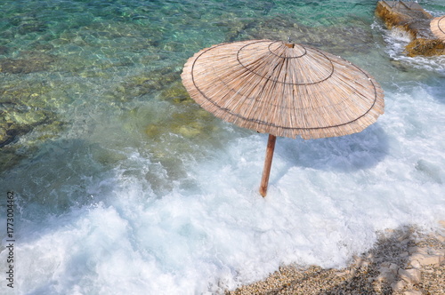 View of golden sand meeting turquoise water under a clear blue sky, framed by a rustic lone thatched parasol. Pebbles beach