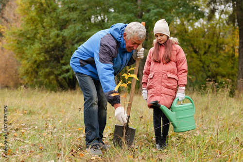An elderly man helps a young girl plant a tree in a lush green field surrounded by colorful autumn leaves. They both wear warm clothing for the cool weather