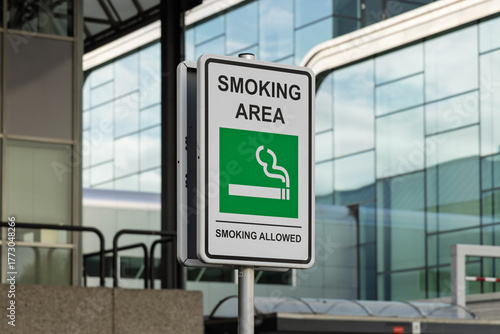 A 'Smoking Area' sign on a pole stands prominently against the backdrop of a modern glass and steel building, confirming that smoking is permitted nearby.