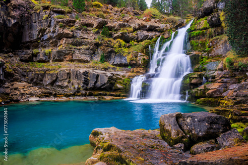 A beautiful waterfall in Ordesa National Park cascades over mossy rocks into a crystal-clear turquoise pool, surrounded by rugged cliffs and lush vegetation in a serene mountain landscape