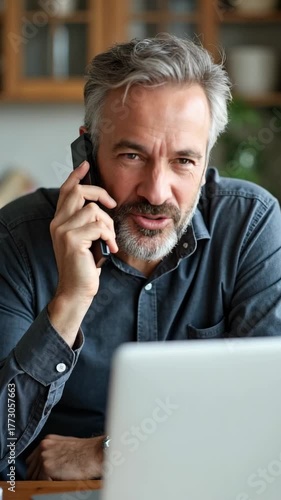 Confident middle-aged man having a phone conversation, sitting at home office desk with laptop, focused on remote work, multitasking and communicating professionally.