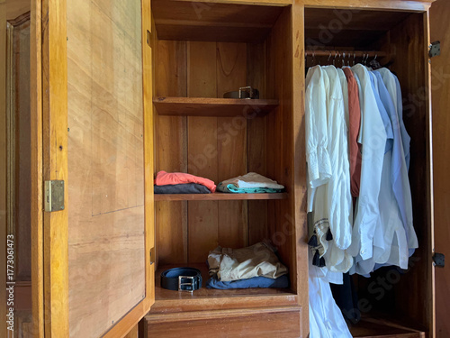 an open wardrobe with a belt on the top shelf and another on the bottom shelf, along with folded clothes and shirts hanging on hangers in the same wardrobe