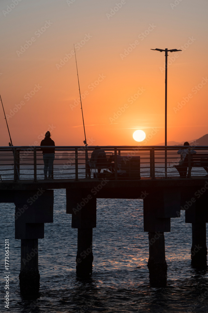 Fototapeta premium Santos city, Brazil. Colorful sunset at Fishermen's Deck. Silhouettes of people fishing and enjoying the view.