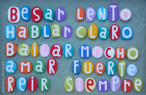 Besar Lento, Hablar Claro, Bailar Mucho, Amar Fuerte y Reir Siempre, creative spanish life slogan composed with hand painted stone letters over green sand 