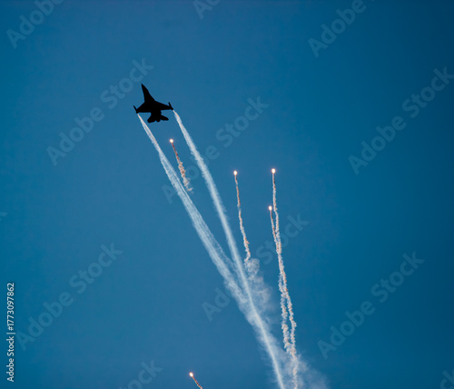 A fighter plane is seen throwing heat flares (flare) in the blue sky background. Bright light traces and smoke clouds reveal the dynamism and visual appeal of the maneuver.