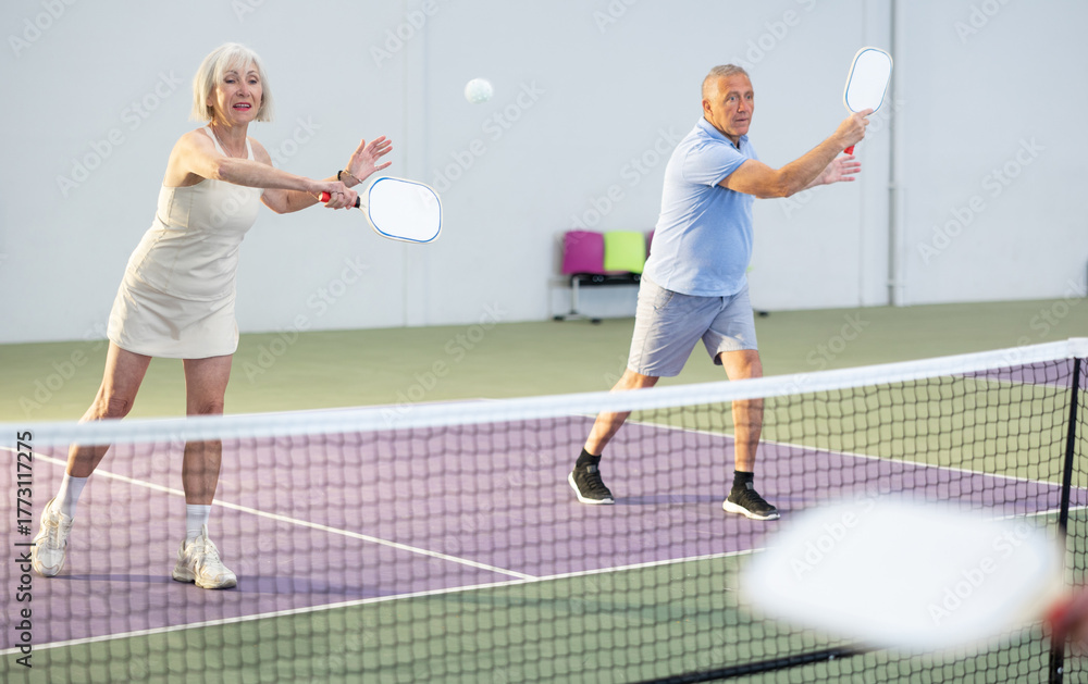 Obraz premium Sporty couple of senior woman and man playing doubles pickleball on indoor court, ready to hit ball. Sport and active lifestyle concept..