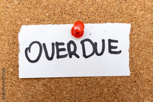 A handwritten note reading OVERDUE pinned to a corkboard, symbolizing overdue tasks, reminders, and deadlines in a workspace environment.