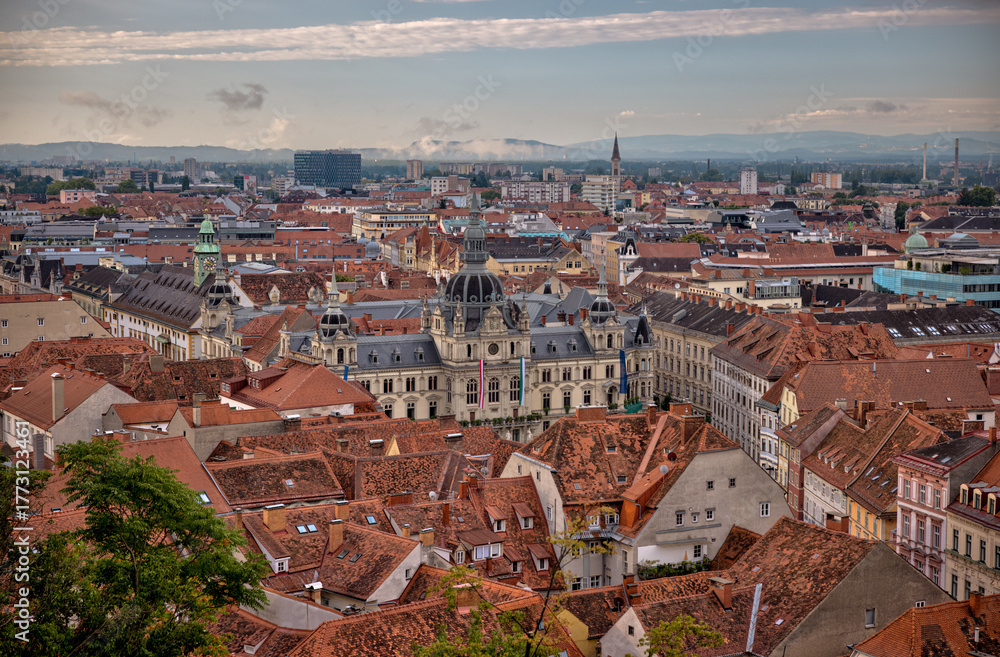 Obraz premium Graz, Austria – July 22, 2025: A scenic panoramic view of the historic city center with red rooftops, church spires, and lush summer foliage under a dramatic morning sky. 