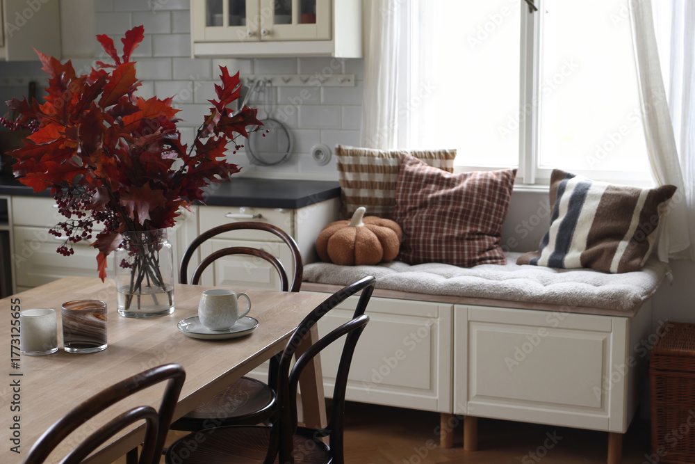 Fototapeta premium Cozy autumn kitchen dininig room interior. Vase of red leaves, candles, cup of coffee tea on table. Window seat with pillows, textile pumpkin. Peaceful stylish Scandinavian home scene, seasonal decor.