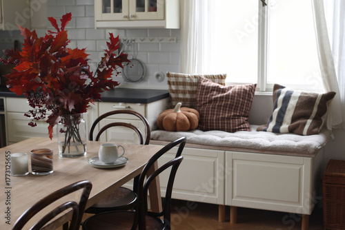 Cozy autumn kitchen dininig room interior. Vase of red leaves, candles, cup of coffee tea on table. Window seat with pillows, textile pumpkin. Peaceful stylish Scandinavian home scene, seasonal decor.