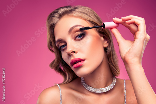Blonde woman applies mascara on pink background showcasing beauty, elegance, and luxury