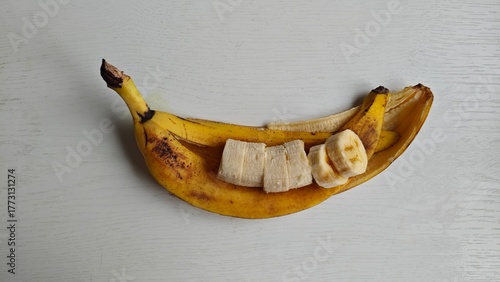 Banana, cut into small circles, lies on peeled skin. Banana slices on ripe peel laid out in shape of smile on white wooden table. Several brown spots on banana peel. Healthy eating, diet