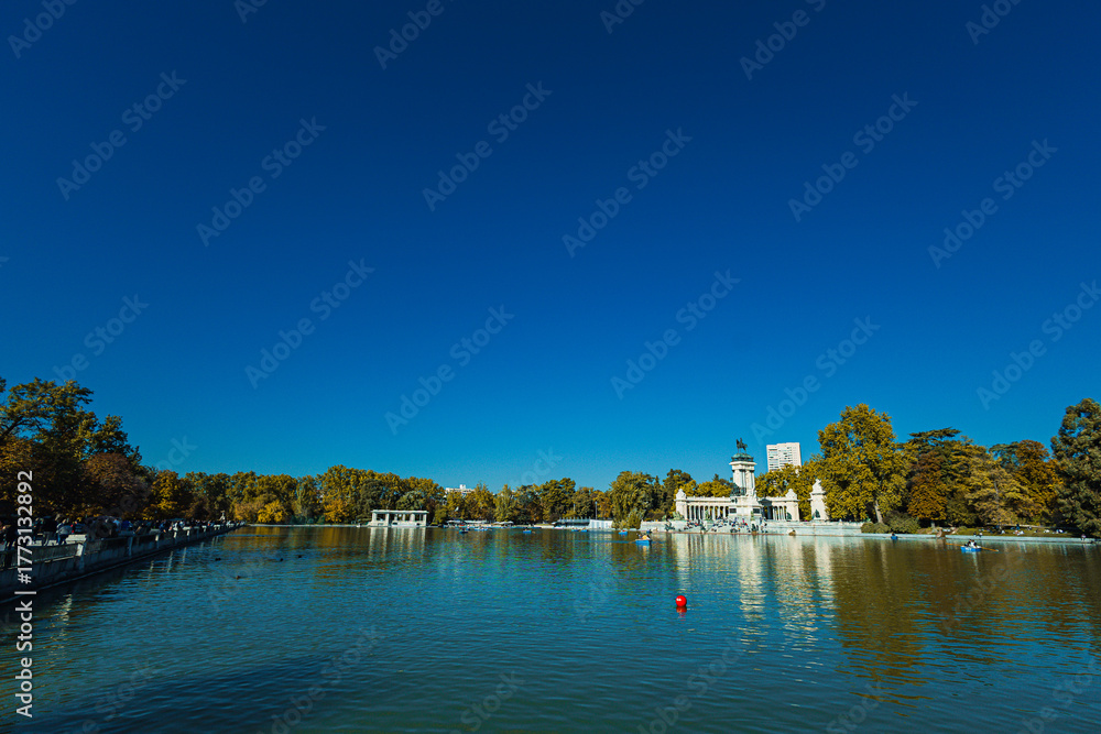 Obraz premium A calm lake reflects the grand monument at El Retiro Park in Madrid. People relax on steps and row small boats under a clear blue sky, surrounded by trees in warm seasonal colors.