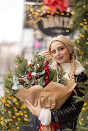 Smiling woman holding a festive Christmas bouquet with fir branches, red berries, and cotton flowers outdoors. Holiday lights and bokeh background create a warm winter atmosphere
