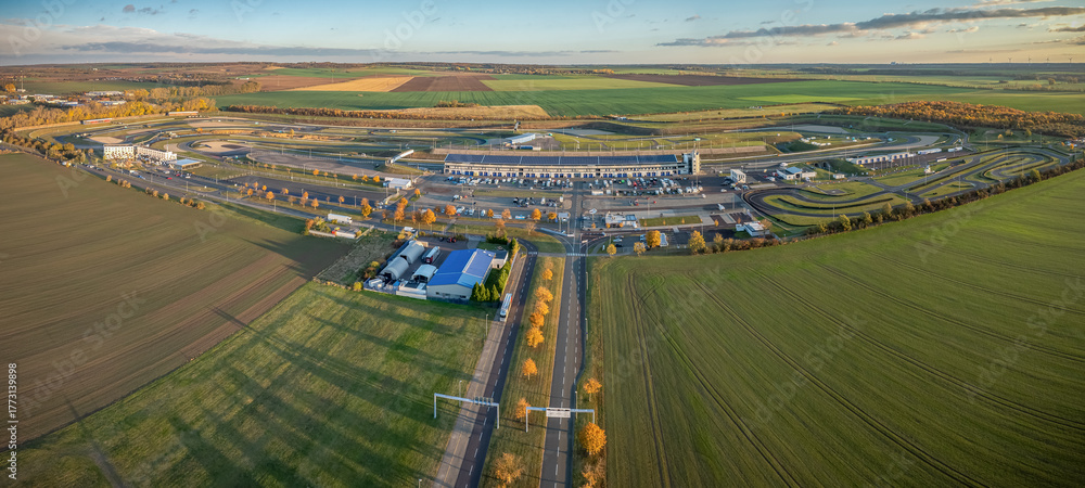 Fototapeta premium Panorama aerial view of large motorsport racetrack complex surrounded by agricultural fields and open countryside. Racetrack with multiple circuits, parking areas, and support buildings.