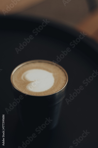 Minimalist coffee aesthetic with creamy latte art in a stylish metal cup on a dark background. Warm tones, soft focus and elegant composition create a cozy, modern cafe vibe. Coffee shop. Hot drink.