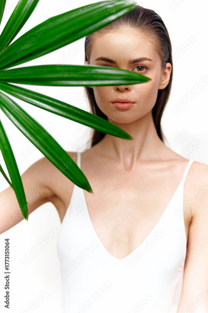 Fototapeta premium Elegant young woman obscured by vibrant green leaf, showcasing serene expression and minimalist style in bright setting. Perfect for lifestyle and beauty themes.