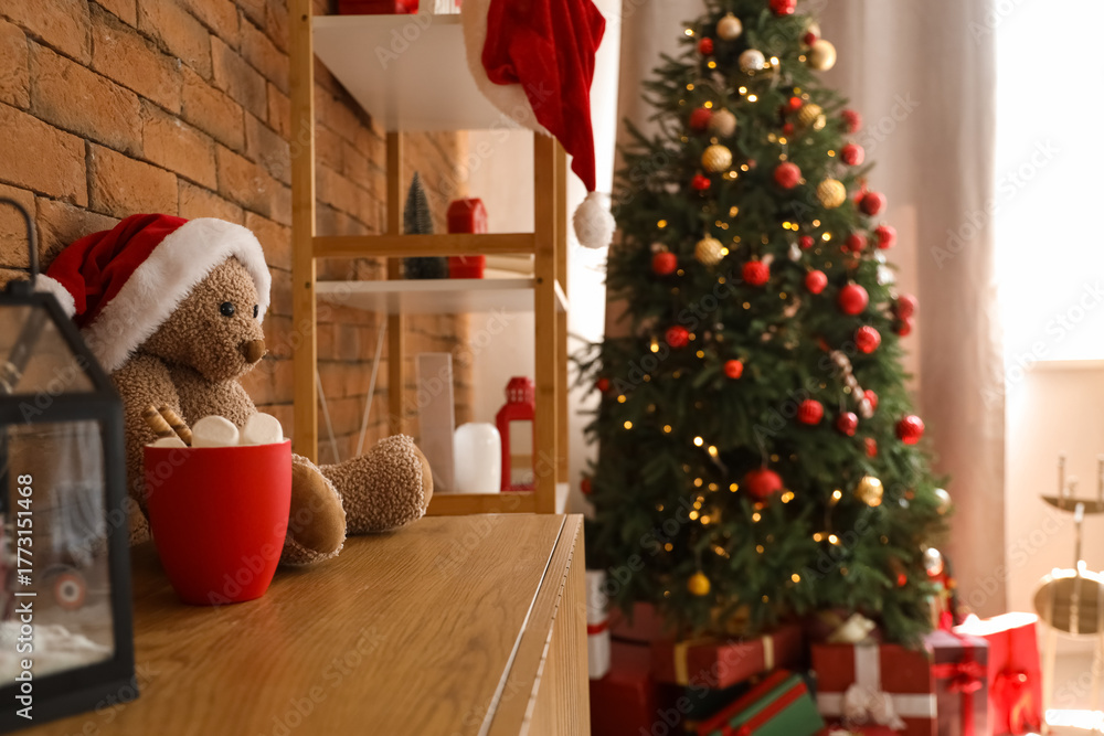 Obraz premium Toy bear with Santa hat and cup of cocoa on drawers in living room decorated for Christmas, closeup