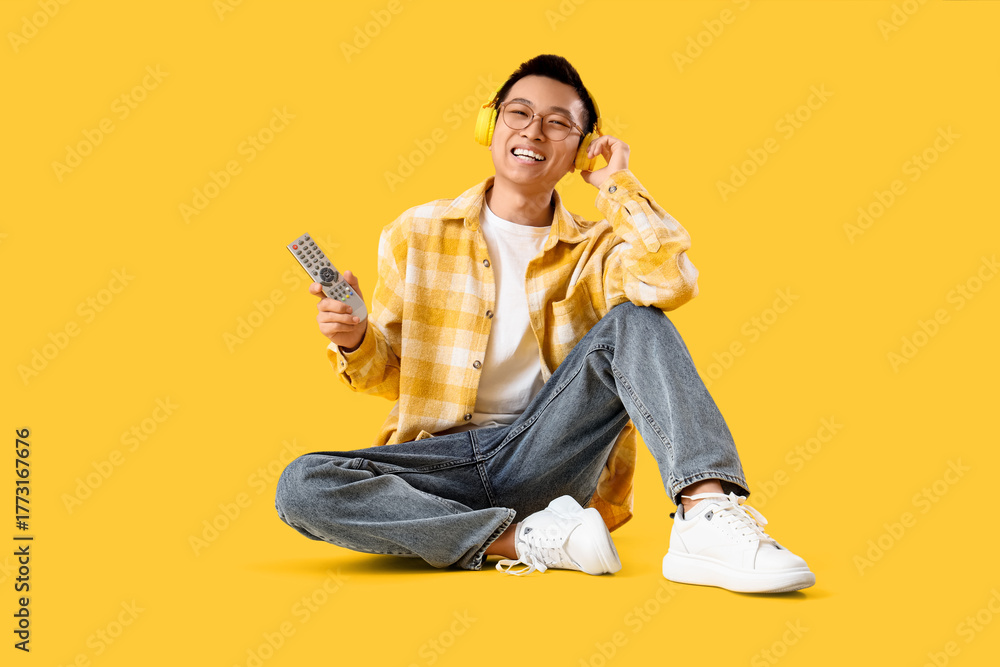 Fototapeta premium Young Asian man with TV remote controller and headphones sitting on yellow background
