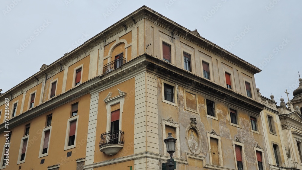 Naklejka premium Rome, Italy - 11 January 2025. Corner of Tempio dei Giovani-Panzieri Fatucci with faded facade, closed red shutters, sculpted window frames, and an upper balcony under overcast daylight.