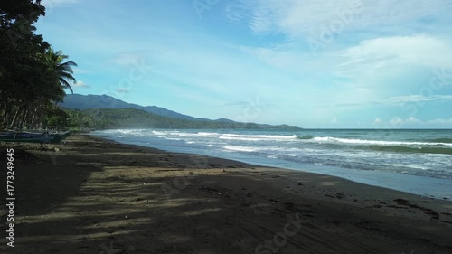 Static shot from shade captures steady waves on slick dark sand, fishing boats resting under coconut palms and distant forested mountains under veiled cloudy sky in Aporawan, Palawan, Philippines.