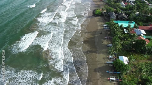 Drone glides low parallel to shoreline showing gentle waves, brightly colored boats shaded by palms and vivid rooftops in dense greenery in Barangay Napsan, Palawan, Philippines.