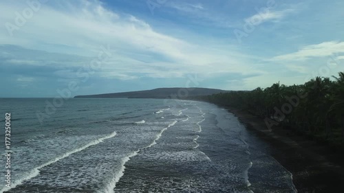 Drone tracks low along extended shoreline with rolling waves, dense palms casting shadows and a distant headland under cloudy morning sky in Barangay Napsan, Palawan, Philippines.