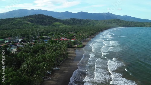 Drone orbits revealing vibrant rooftops nestled in dense palm canopy along curving shoreline with steady waves and blue-hued mountains rising in the background in Barangay Napsan, Palawan, Philippines