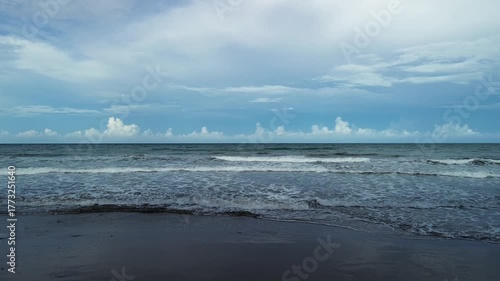 Camera slowly pulls back from the shoreline at Little Siargao in Aporawan, Aborlan, Palawan, revealing even waves on dark sand, vast ocean, and soft clouds with distant cumulus formations.