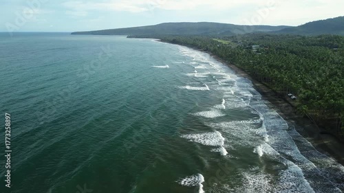 Drone flies forward at altitude showing emerald waters with deliberate waves on sand, wall of coconut palms casting shadows and distant headland in Barangay Napsan, Palawan, Philippines.