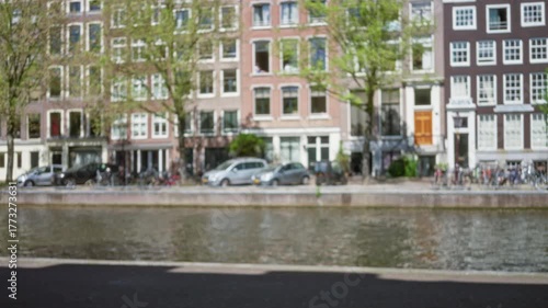 Blurred old buildings and canal in amsterdam on a sunny day with bicycles and parked cars lining the waterway and green trees adding to the scenic streetscape.