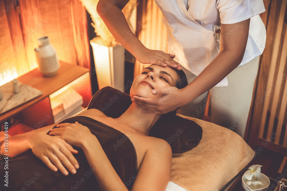 Fototapeta premium Caucasian woman enjoying relaxing anti-stress head massage and pampering facial beauty skin recreation leisure in warm candle lighting ambient salon spa in luxury resort or hotel. Quiescent
