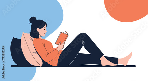 A young woman with dark hair tied in a bun is comfortably reading a book while lying on her side against pillows, enjoying a moment of leisure.