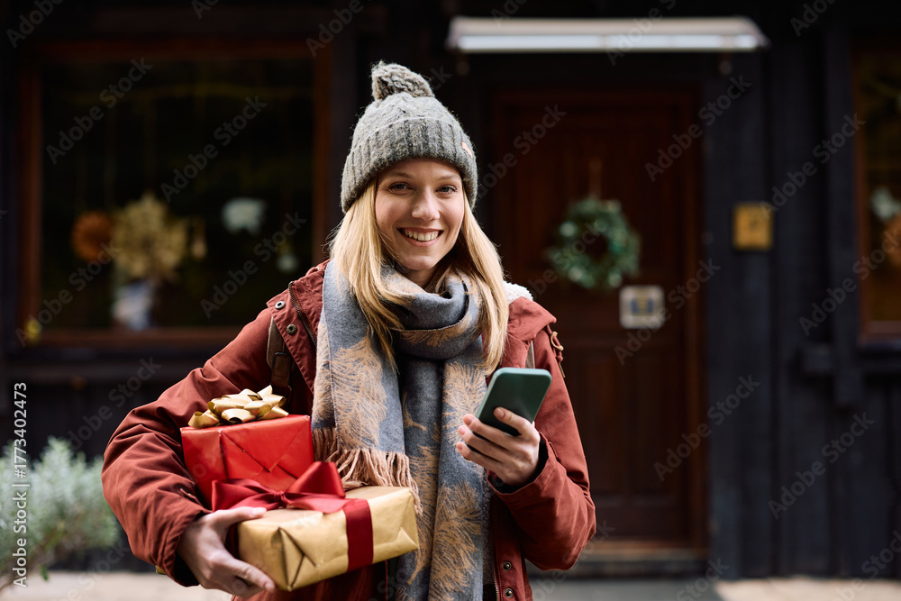 Fototapeta premium Happy woman using cell phone while holding Christmas presents on front of her house.