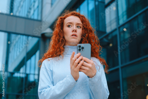 Young woman with red curly hair looks surprised while using her smartphone outside modern building