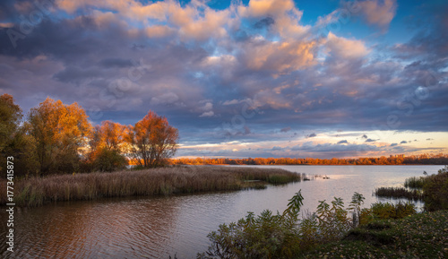 Sunset over the Dnipro River on Obolon Island in Kyiv, with autumn trees glowing in golden light under dramatic clouds.
