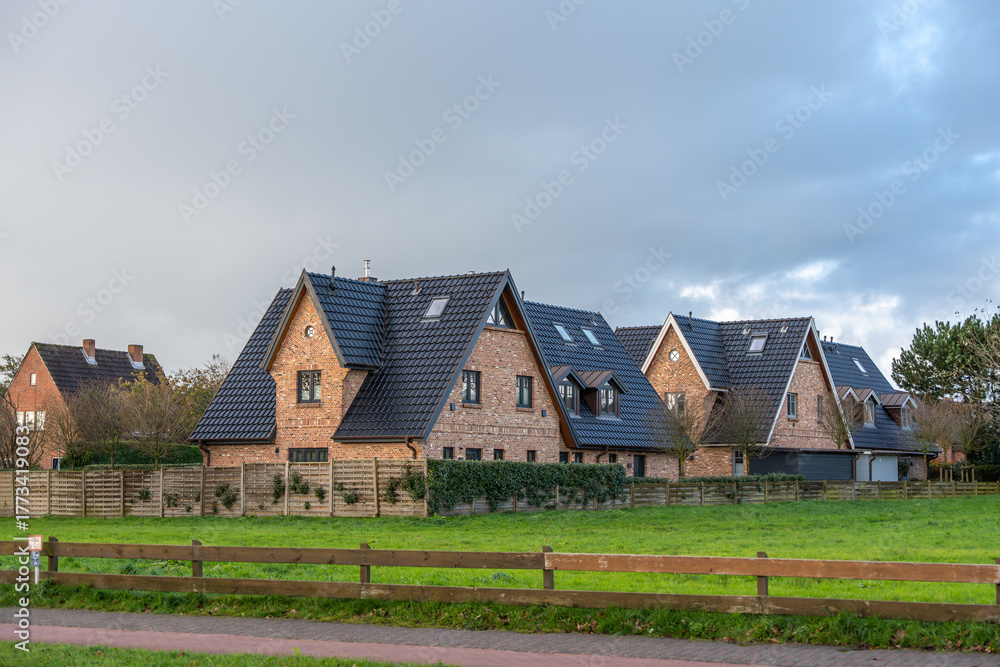 Fototapeta premium Residential brick houses with dark roofs. Modern brick houses with steep dark roofs in a quiet suburban neighborhood. The view shows fenced gardens and green lawns under a cloudy sky.
