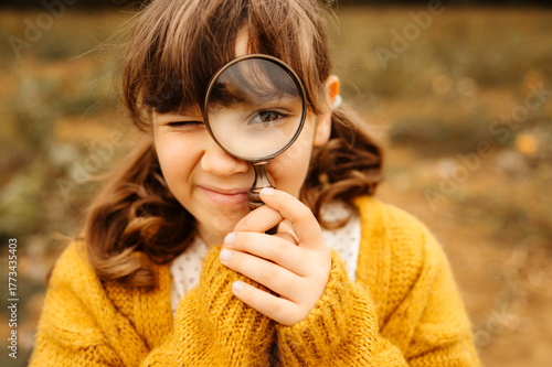 Girl in yellow sweater looking through magnifying glass outdoors.