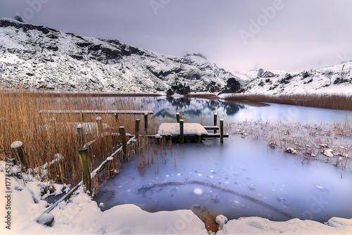 Snow covered landscape at La Arboleda in Bizkaia, Basque Country. Frozen lake with wooden pier and reeds surrounded by mountains under a cold winter atmosphere