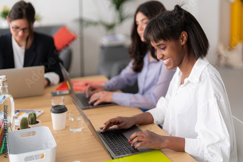 Three diverse women collaborate in a modern office, each focused on their laptops. The smiling Black woman in the foreground actively types, alongside her concentrating colleagues.