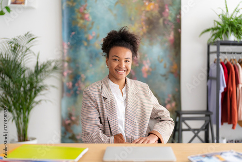 A smiling young Black woman sits at a desk in a bright office. She wears a plaid blazer, looking confidently at the camera. Her workspace includes a laptop and colorful folders.