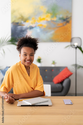 A smiling young Black woman sits at a desk in a modern home office. She has a laptop, notebook, and phone on the wooden surface, ready for work or study.