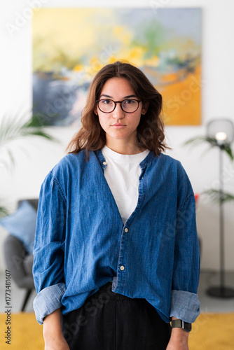 A young woman with long brown hair and glasses stands confidently indoors, looking directly at the camera. She wears a blue denim shirt over a white t-shirt, portraying a casual yet focused demeanor.