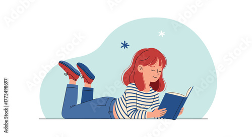 A young woman with red hair is comfortably lying on her stomach, engrossed in reading a blue book, enjoying a moment of quiet relaxation.