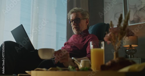 Gray Haired Man Sits Relaxed in Hotel Chair, Sipping Coffee and Typing on Laptop in Morning Light and Cozy Atmosphere. Concept of Remote Work, Business Travel, and Digital Flexibility. Handheld.